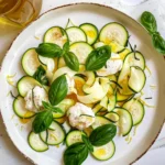 Fresh Zucchini Crudo with Burrata on a white plate, topped with creamy cheese, bright green basil leaves, and drizzled olive oil, artfully arranged for a vibrant summer appetizer.